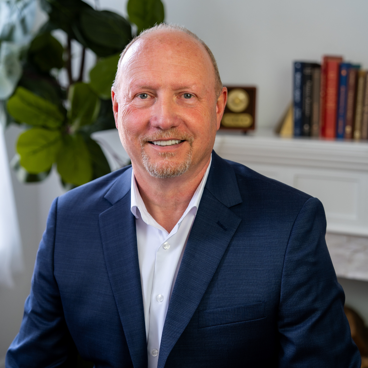 Headshot of Financial Advisor in Navy Blue Blazer with a backdrop of an office featuring a fig tree and bookshelf